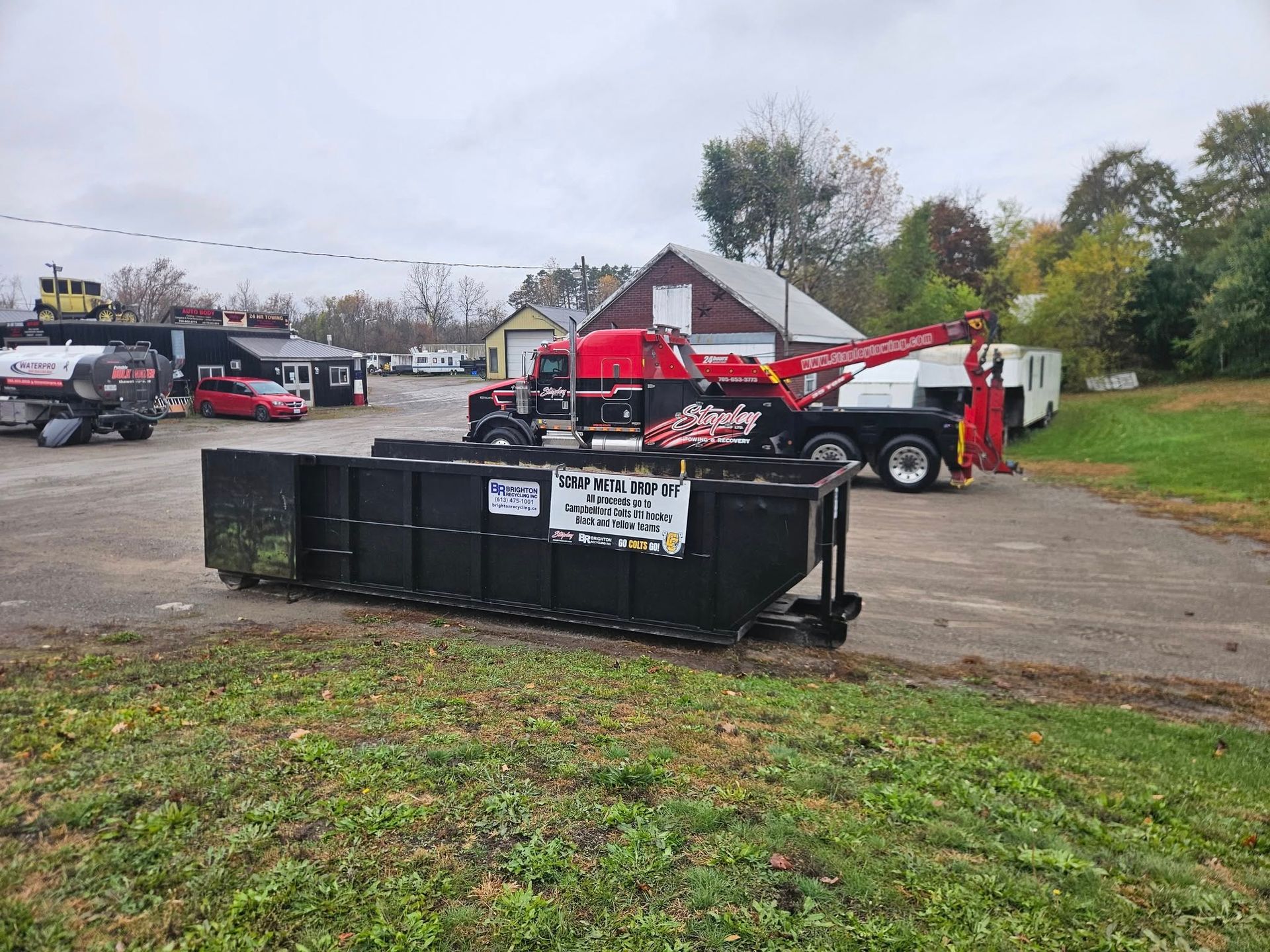 Red tow truck loading a black dumpster in a parking lot. Buildings and trees in background. Overcast sky.