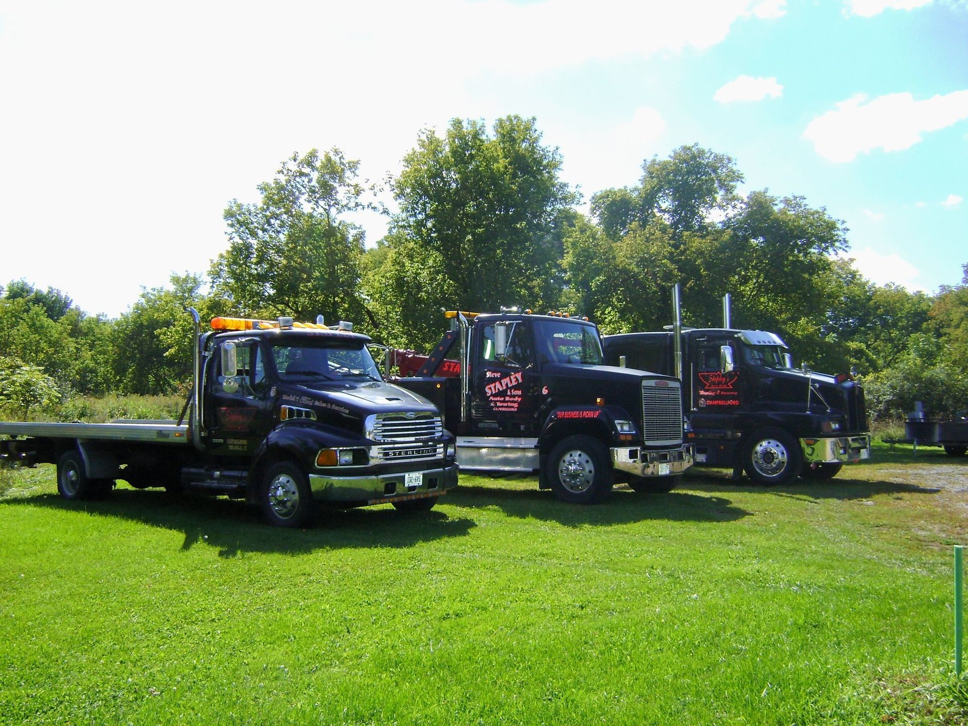 Three black tow trucks parked on a grassy area, trees in the background under a bright sky.