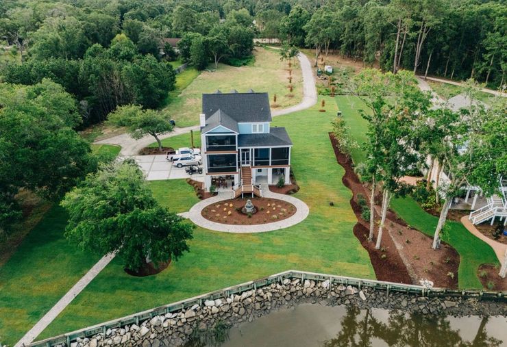 Aerial view of a two-story blue house with porch, circular driveway, and well-manicured lawn near water and trees.