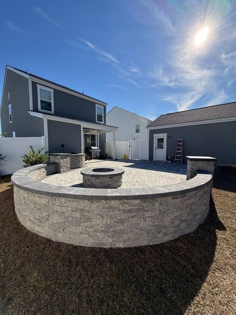 A circular brick patio with fire pit, surrounded by a low wall, in a backyard. Houses and blue sky in background.