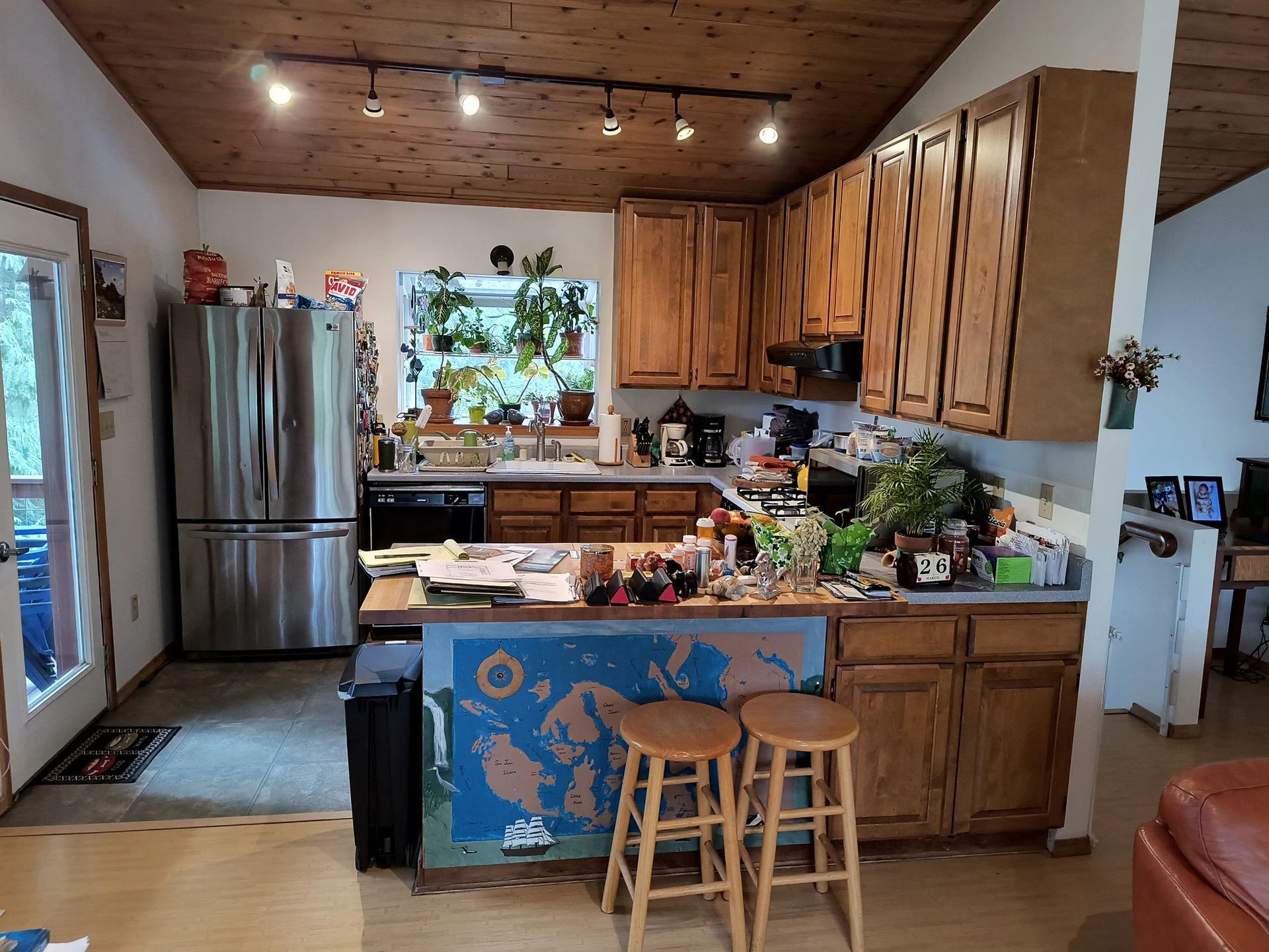 Kitchen with wooden cabinets, stainless fridge, central island, and two stools under warm ceiling lights.
