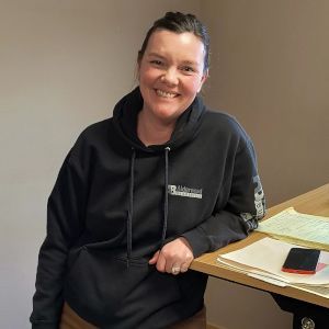 Smiling person in a black hoodie seated at a desk with papers and a phone