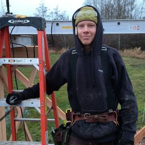 Person in a black hoodie and safety harness standing on a ladder outdoors, holding a tool belt.