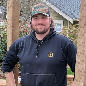 Smiling person in a black hoodie and camo cap standing on a wooden deck outdoors