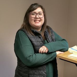 Smiling person in glasses leans on a desk, wearing a green shirt and black vest.