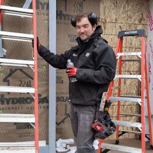 Worker in black jacket holding a drink, standing between two ladders at a building under construction.