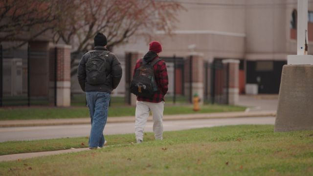 Two people with backpacks walk side-by-side on a sidewalk near a lawn and buildings on an overcast day.