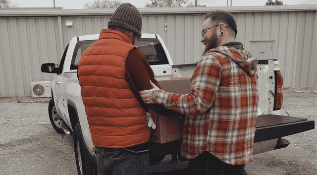 Two people in casual outdoor clothing unload a large cardboard box from the bed of a white pickup truck.