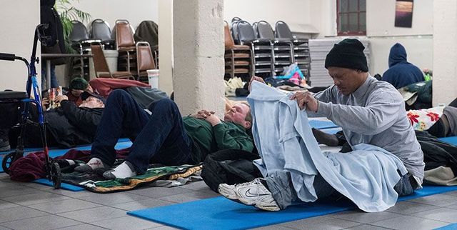 People resting on mats on the floor in a large indoor shelter, with one person folding a light blue blanket.