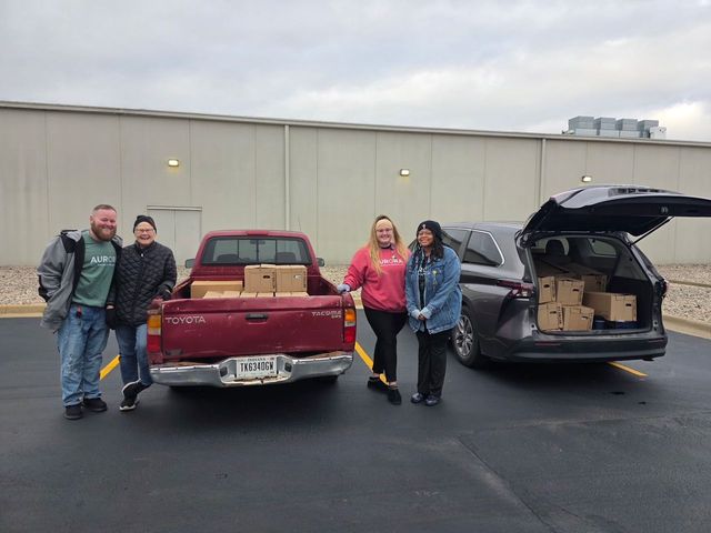 Four people stand by a red pickup truck and a dark SUV loaded with cardboard boxes in an outdoor parking lot.