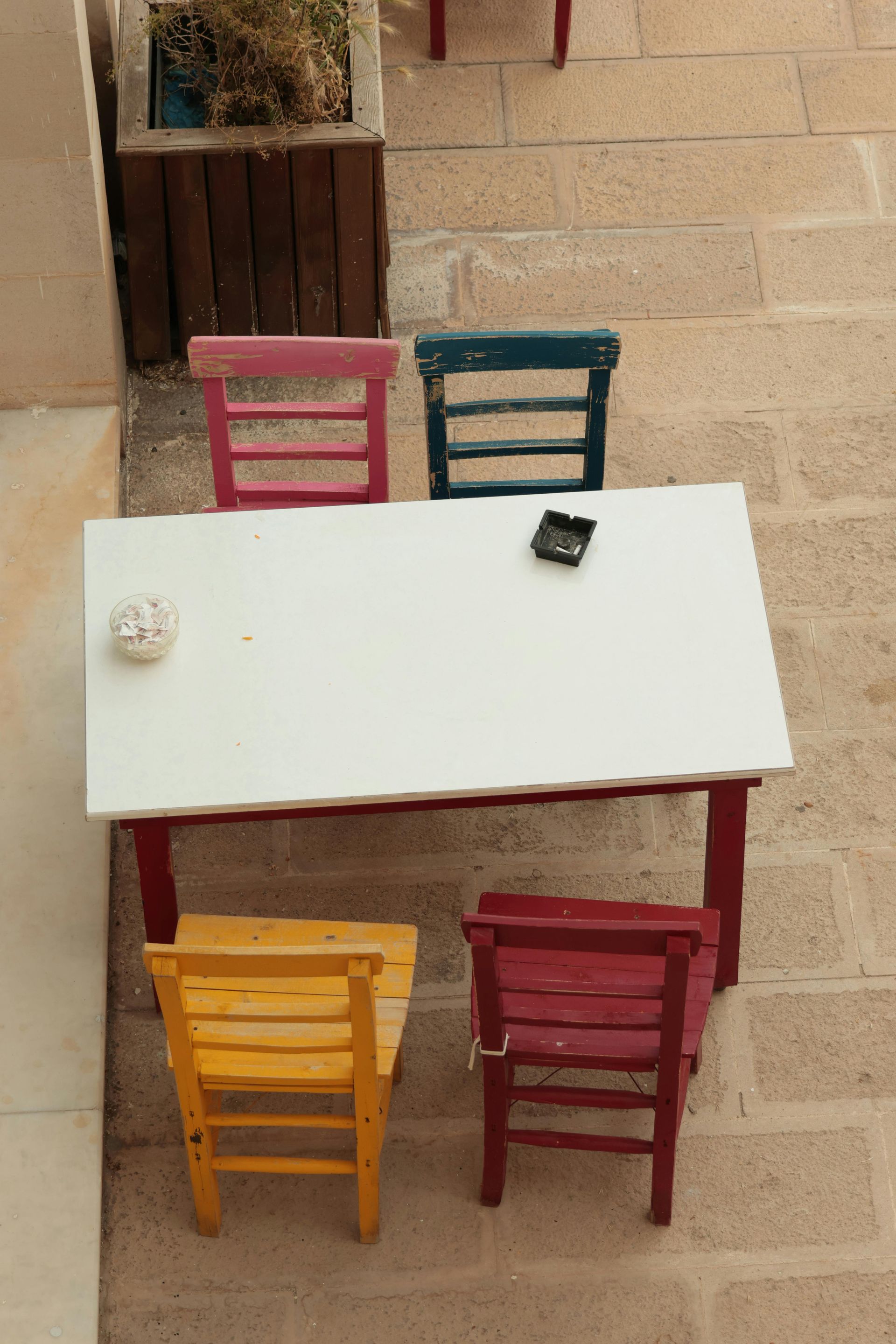 Sillas de colores alrededor de una mesa blanca en un patio de ladrillo. Un pequeño cenicero y un cuenco reposan sobre la mesa.