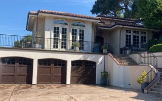 Cream-colored house with three-car garage, upper balcony, and arched French doors. Brown garage doors and stairs leading up.
