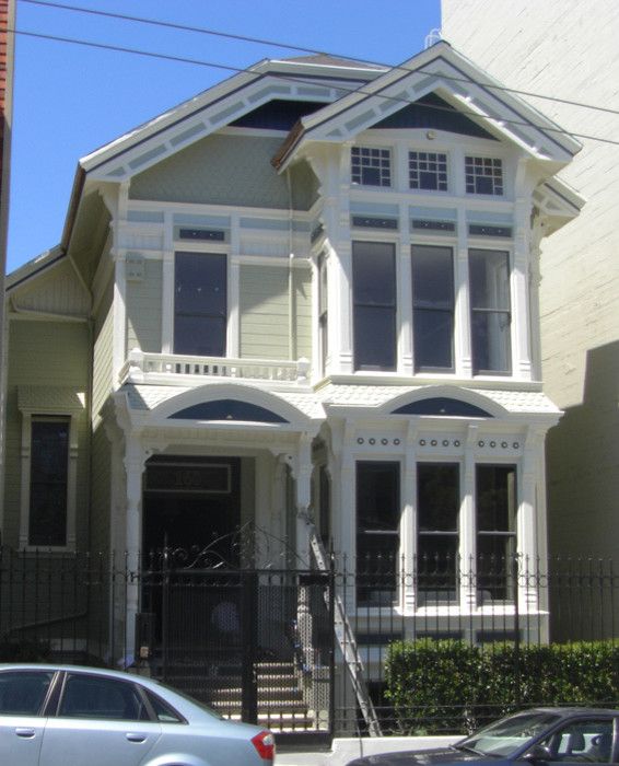 Victorian style house with light green siding and white trim.  Two stories with a front porch, black fence, and cars parked in front.