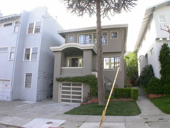 Two-story gray house with garage, windows, and small front yard; flanked by other houses on a sunny day.