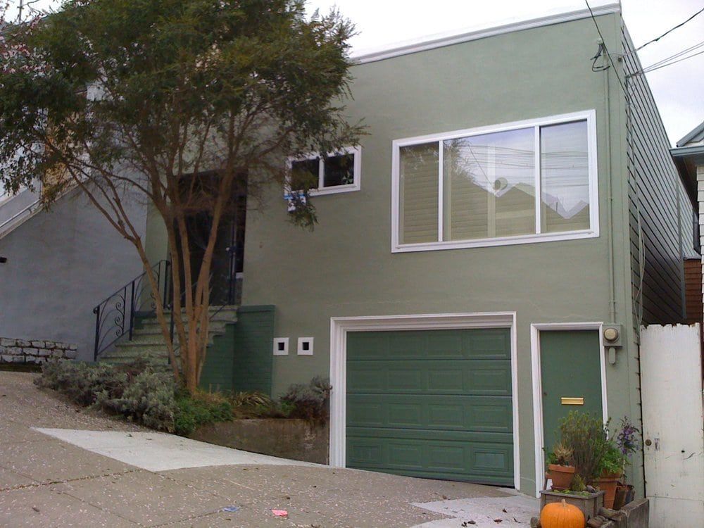 Green two-story house with green garage door, front steps, and tree in front on a sloping street.