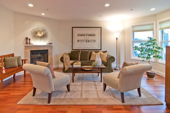 Living room with brown wooden floor, olive green sofa, beige armchairs, fireplace, and large windows.