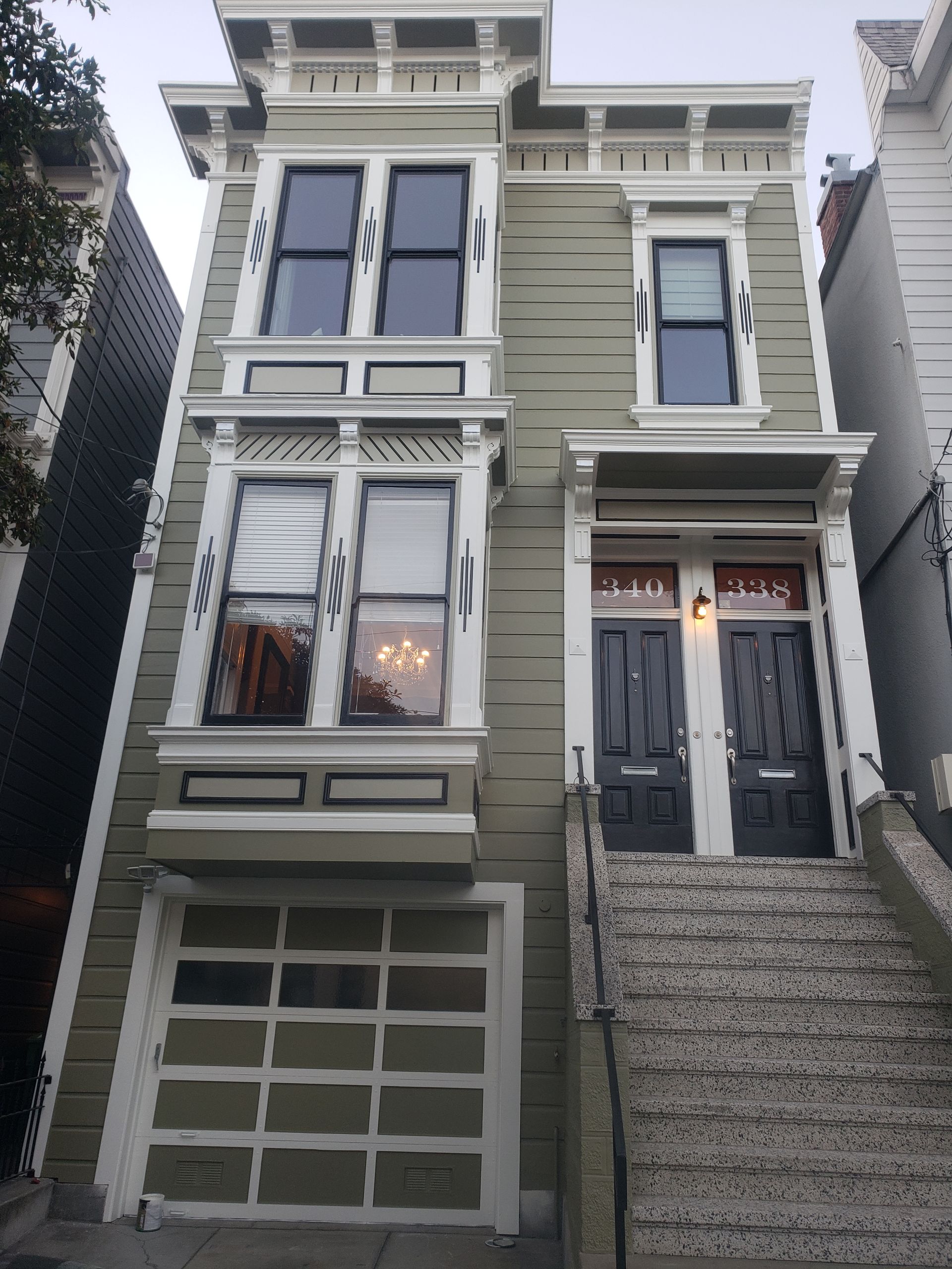 Multi-story house with green siding, bay window, two black front doors, and garage. Stone steps lead up to doors.