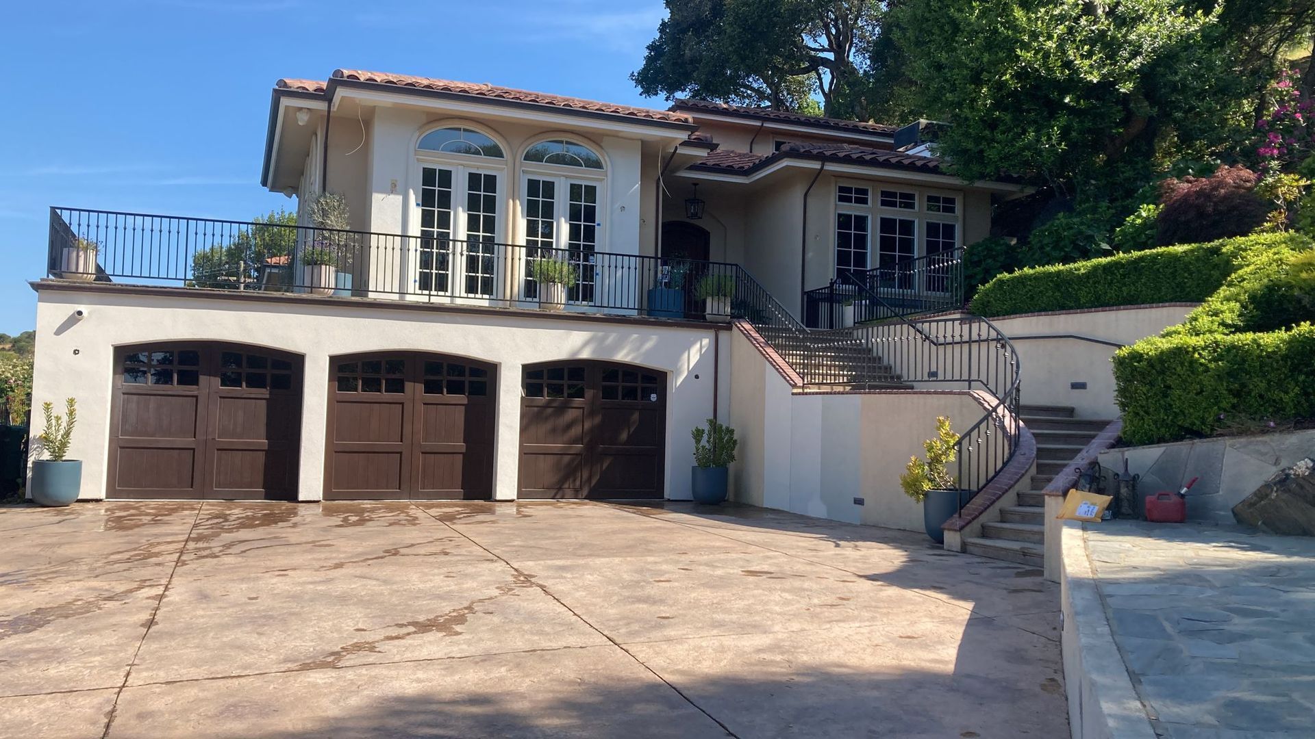 Mediterranean-style house with three-car garage, upper balcony, and staircase leading to entrance. Brown garage doors, tan exterior, blue sky.