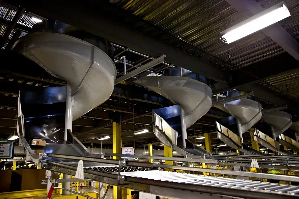 Automated warehouse conveyor system with chutes, lit by overhead fluorescent lights. Talos Engineered Products