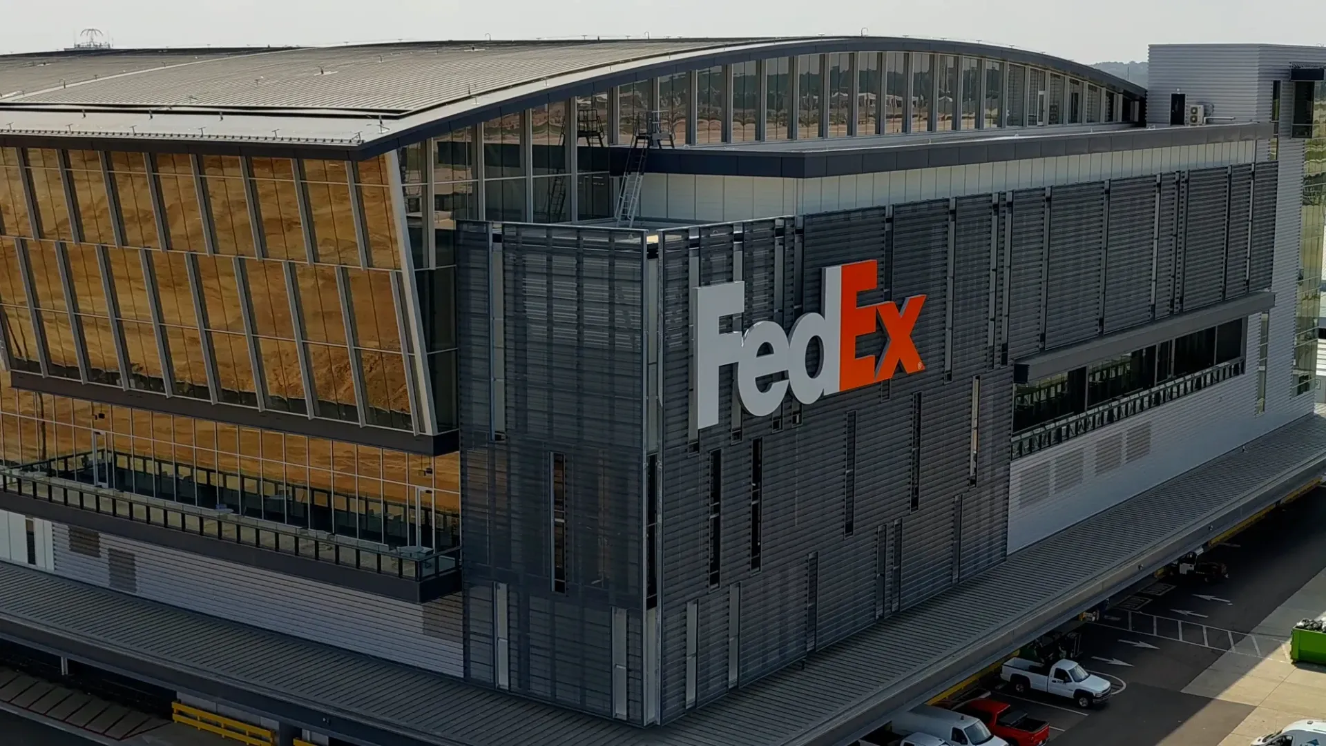 FedEx building with a large orange logo. Exterior shot. Gray and brown facade with parked vehicles.