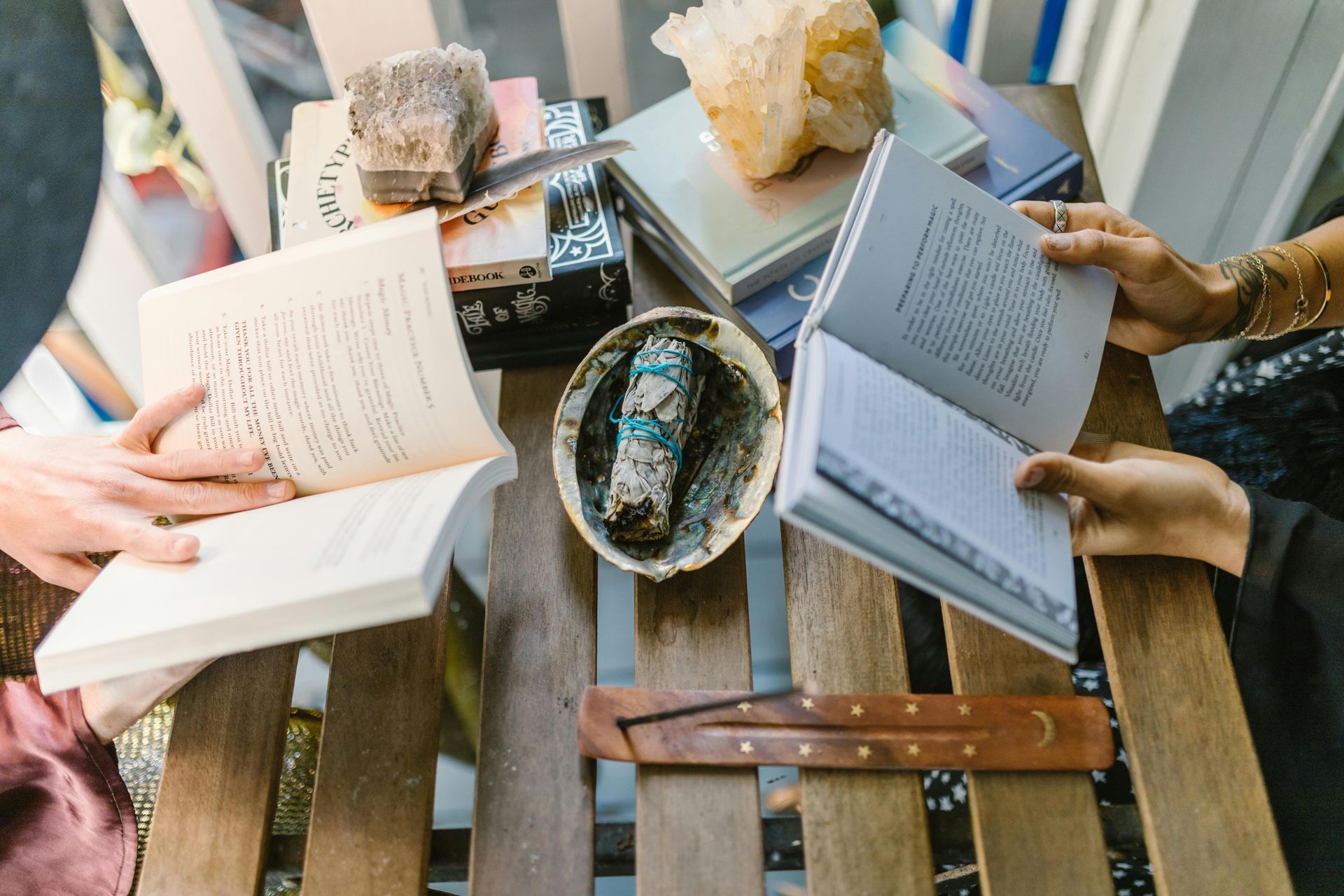 Picture of two people reading books, by piles of books held down by crystals, with sage in a bowl in between them