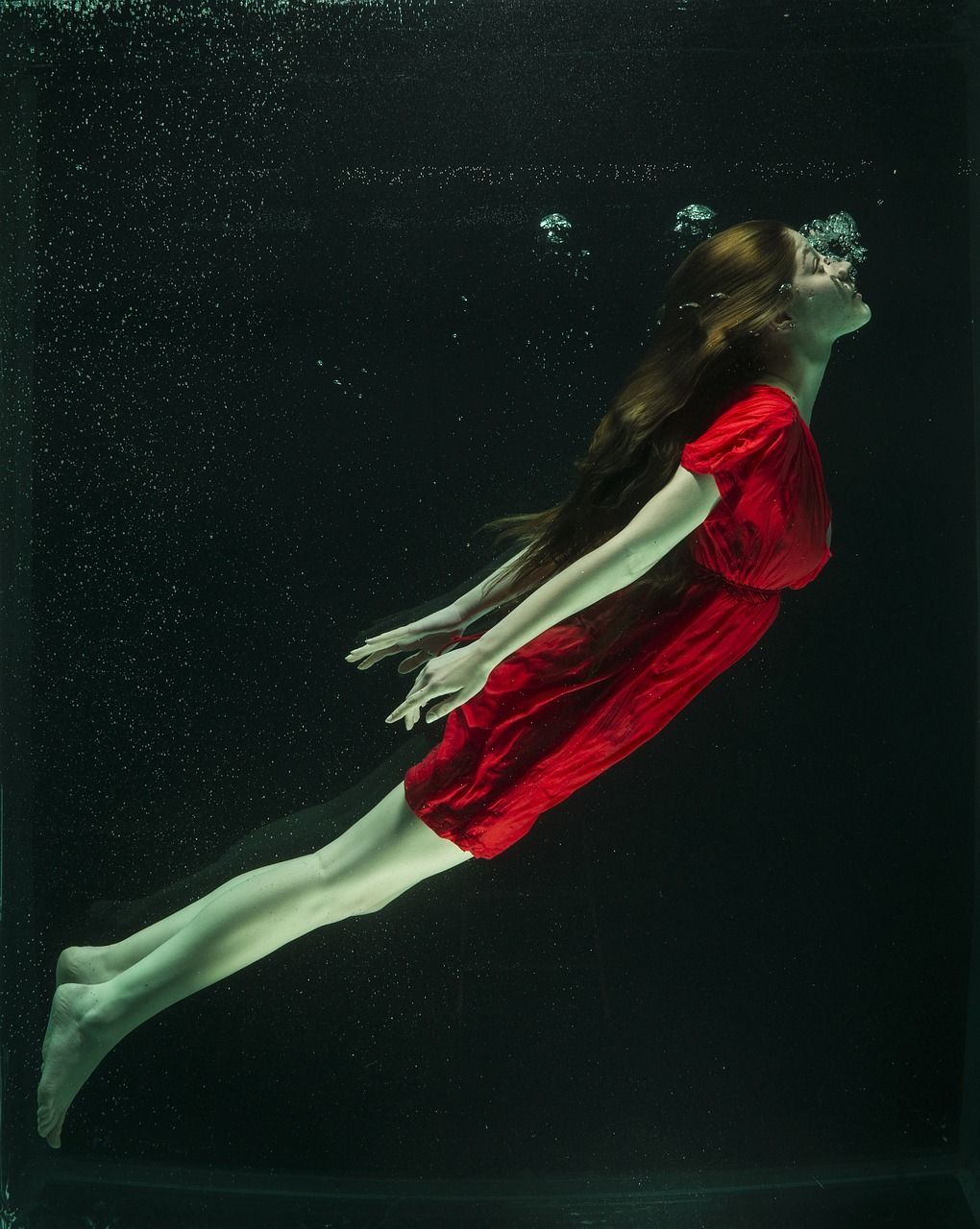 Photo shows Girl in a red dress, under water, blowing bubbles of air as she dives back up towards the surface