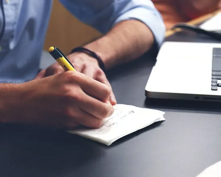 A man is writing on a piece of paper next to a laptop