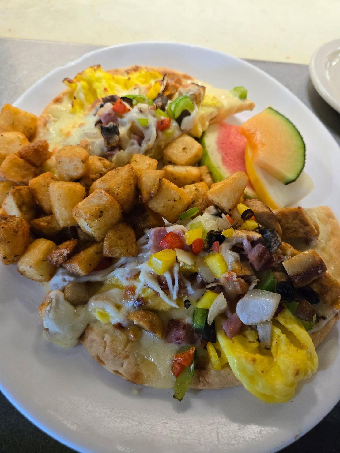Deux pains pita pour le petit-déjeuner, garnis d'œufs, de poivrons, d'oignons et de viande, servis avec des pommes de terre rissolées et des tranches de fruits.