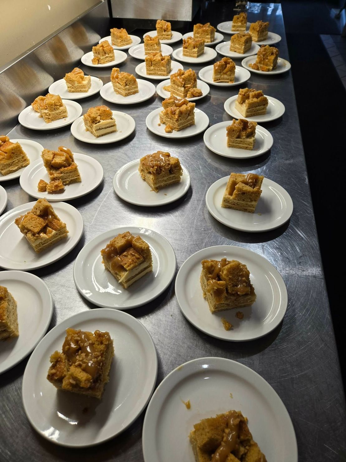 De nombreuses petites assiettes surmontées de morceaux de gâteau carrés sont disposées sur un long plan de travail métallique de cuisine professionnelle.