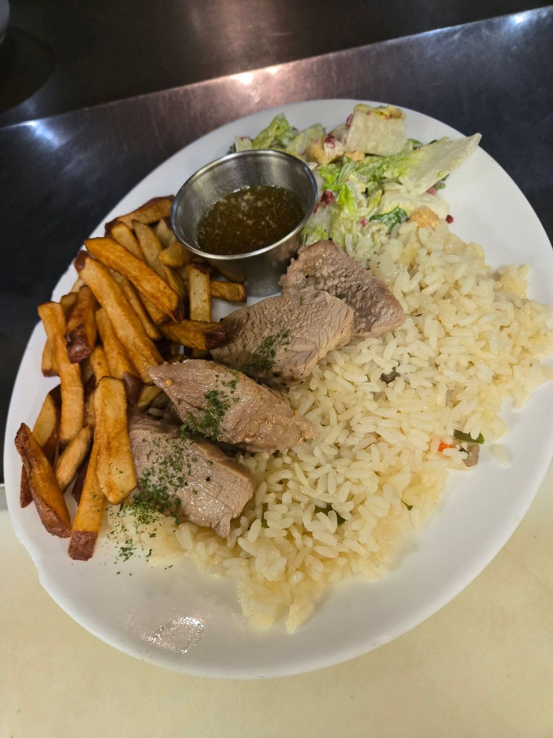 Une assiette composée de viande assaisonnée, de riz, de frites, d'une petite coupelle de sauce brune et d'une salade d'accompagnement.