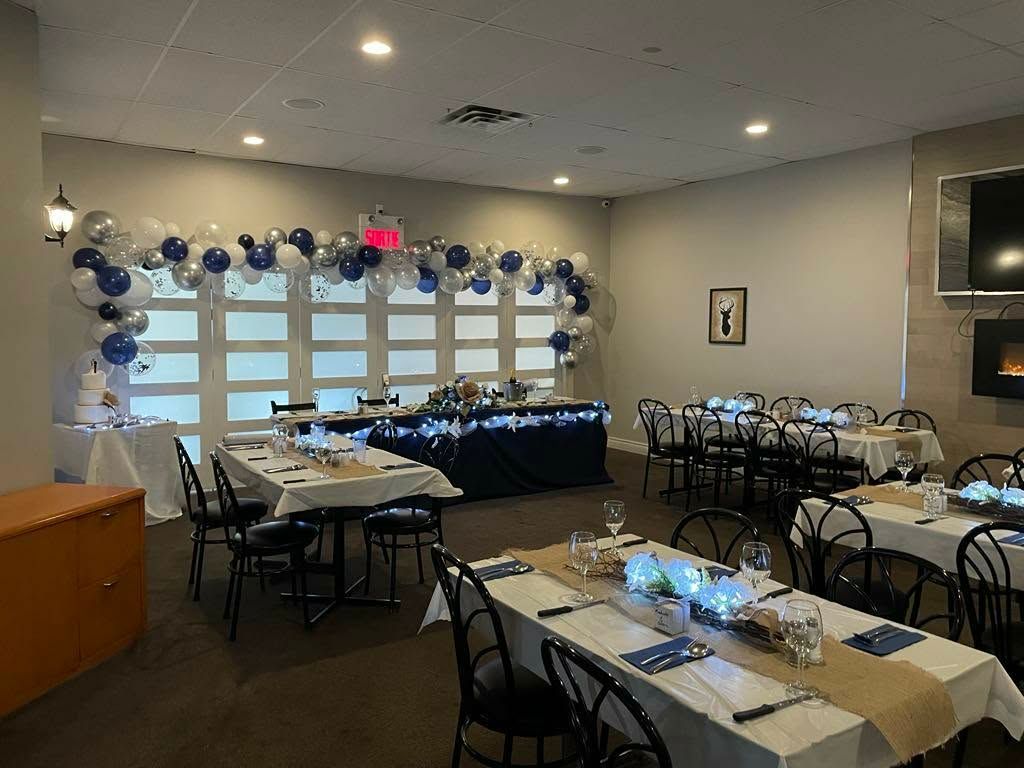 Une salle de banquet aménagée pour une fête avec des arches de ballons bleus et blancs, des tables décorées et un buffet recouvert d'une nappe sombre.