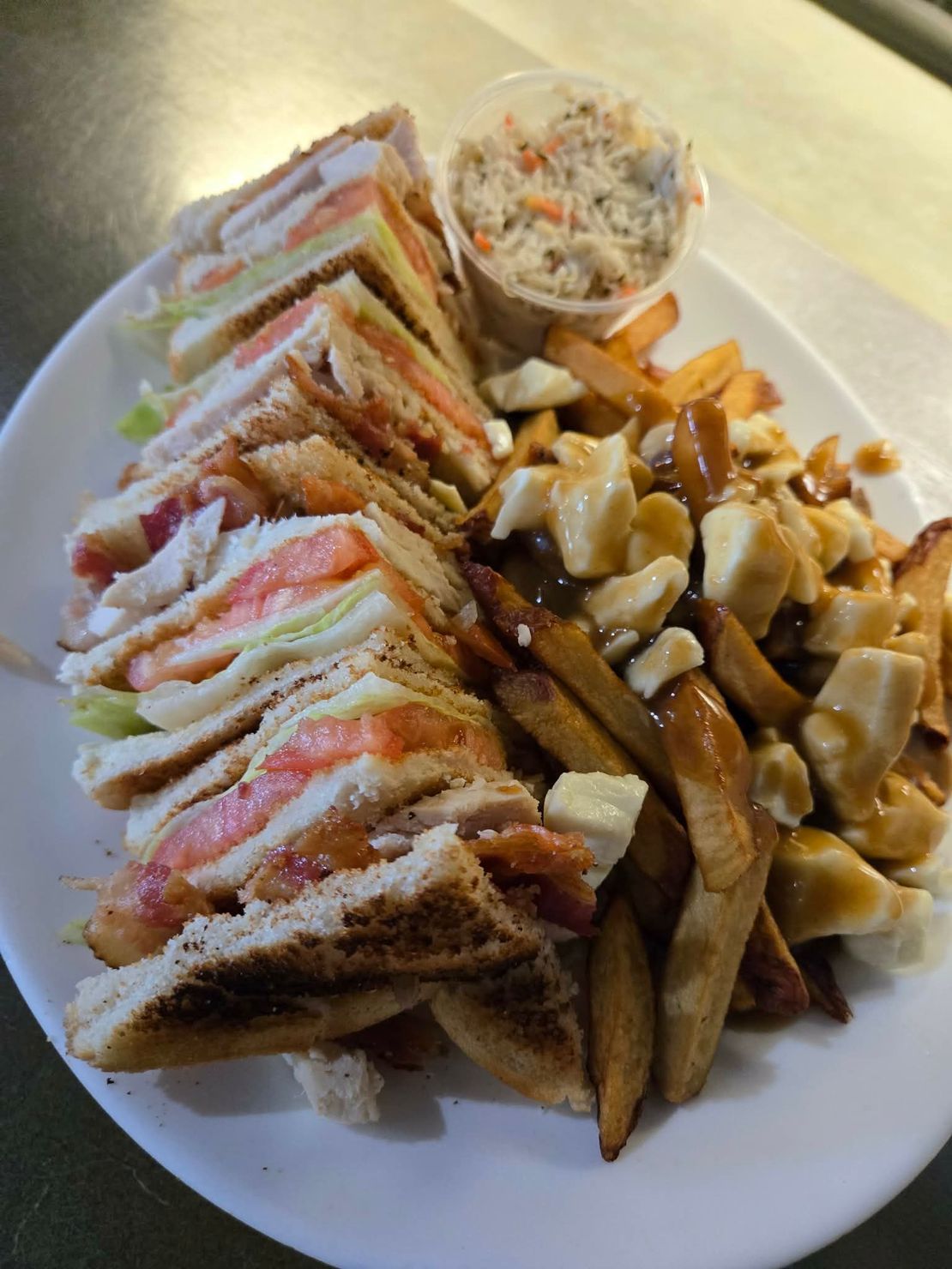 Un club sandwich coupé en quatre, une portion de poutine avec sauce et fromage en grains, et une petite tasse de salade de chou dans une assiette.