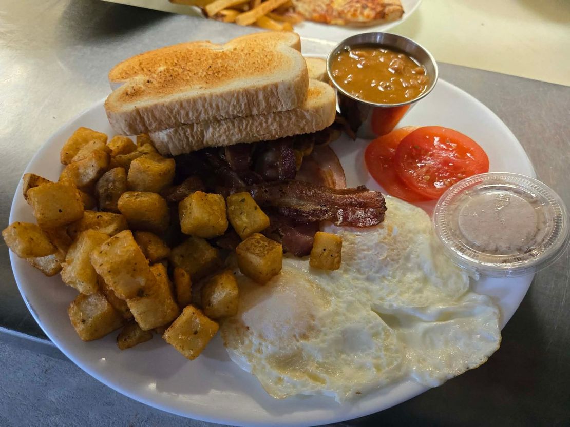 Une assiette de petit-déjeuner avec des œufs, du bacon, des pommes de terre rissolées, du pain grillé, deux tranches de tomate et une petite coupelle de sauce, posée sur une table.