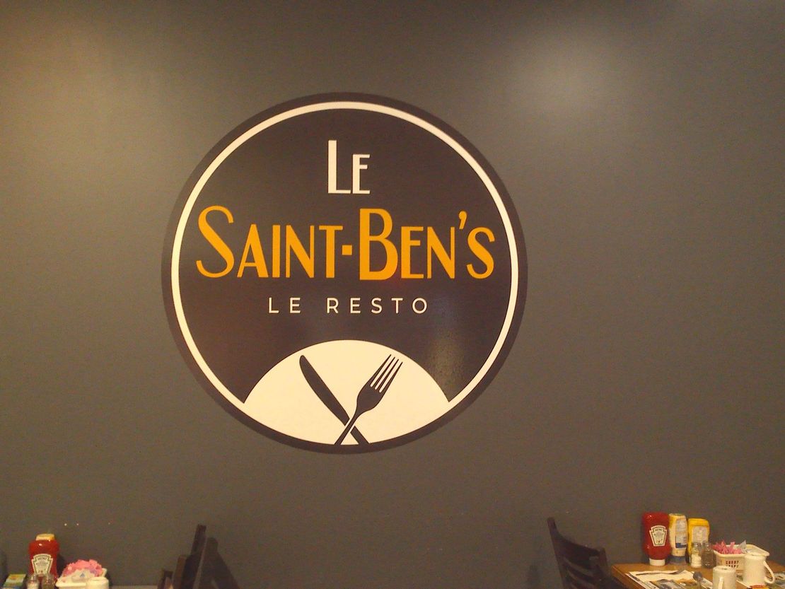 Une enseigne circulaire pour « Le Saint-Ben's Le Resto » avec un texte doré et une icône blanche représentant un couteau et une fourchette sur un mur sombre.