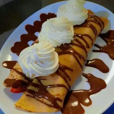 Une crêpe roulée garnie de trois volutes de crème fouettée et nappée de sauce au chocolat, servie sur une assiette blanche.