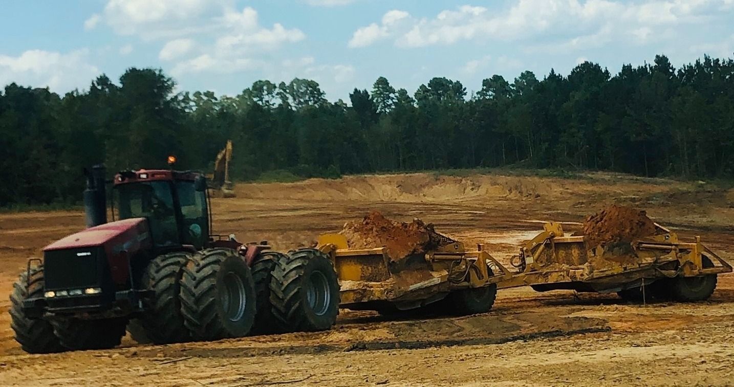 Red tractor pulling a large dirt scraper across a muddy construction site with trees in the background