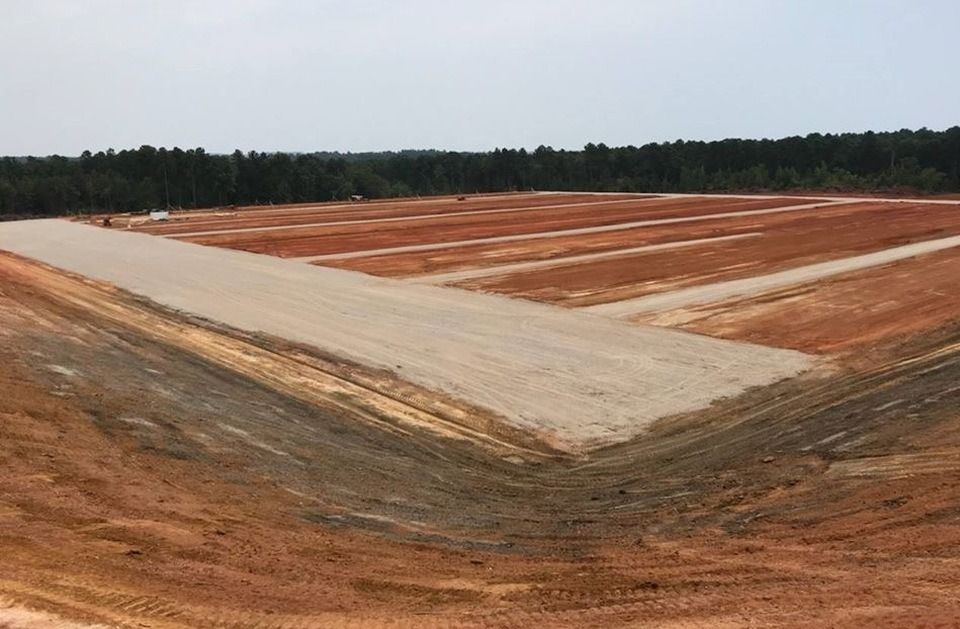 Cleared construction site with red soil, a wide pale dirt road, and forest in the distance