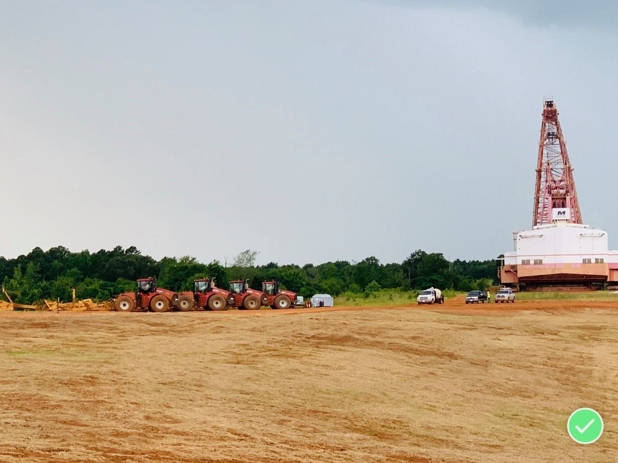 Open field with a drilling rig on the right and vehicles near trees under an overcast sky.