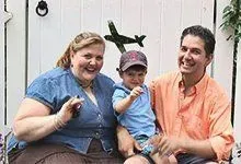 Three people sitting outdoors, smiling, with a child in a cap between two adults