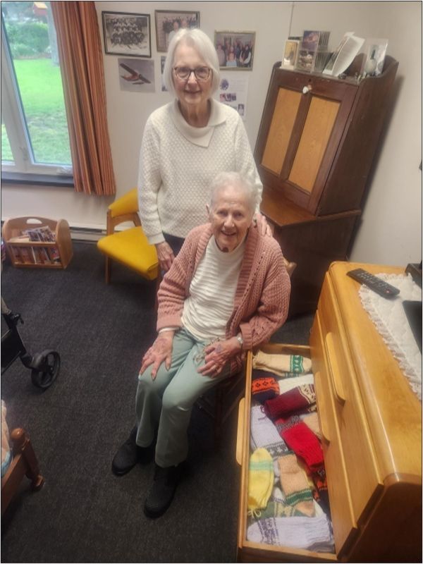 Two elderly women smiling in a cozy room, one seated and one standing beside her.