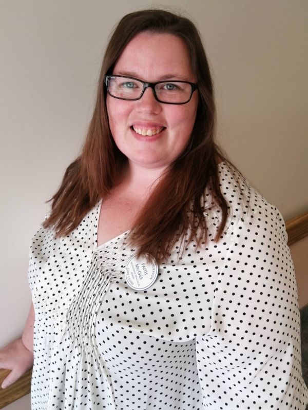 Smiling person in glasses wearing a white polka-dot blouse against a plain wall