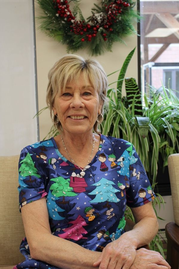 Smiling woman in colorful Christmas shirt seated indoors beside a wreath and potted plant.