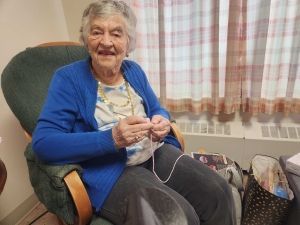 Smiling elderly woman in a blue cardigan sitting in a chair, knitting in a cozy room