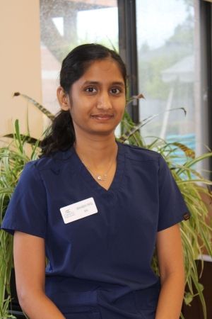Person in navy scrubs with a name badge, standing indoors in front of plants and a window.