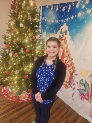 Woman standing beside a lit Christmas tree and festive holiday backdrop.