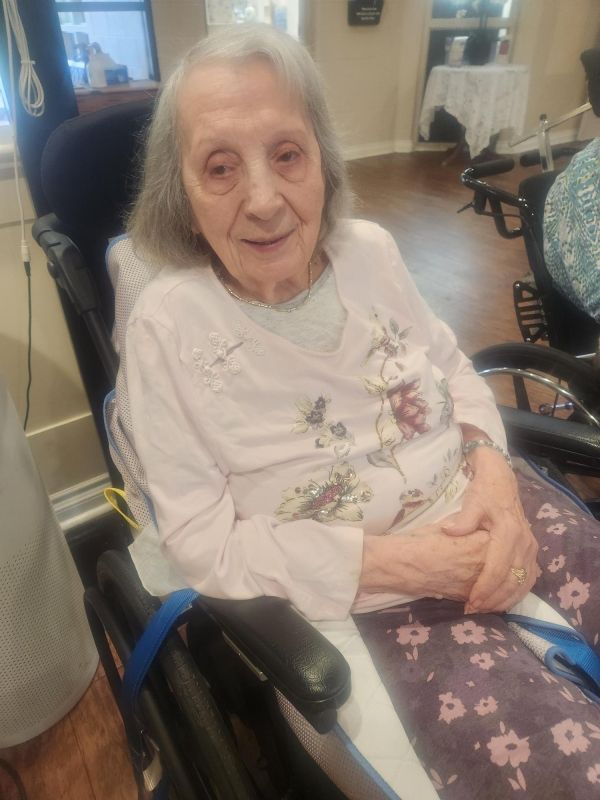 Elderly woman seated in a wheelchair, wearing a light pink top, in a home setting.
