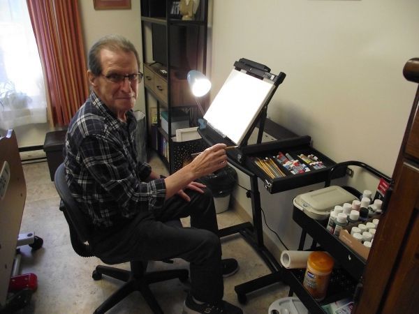 Man seated at an easel painting, with art supplies and bright light in a small studio.