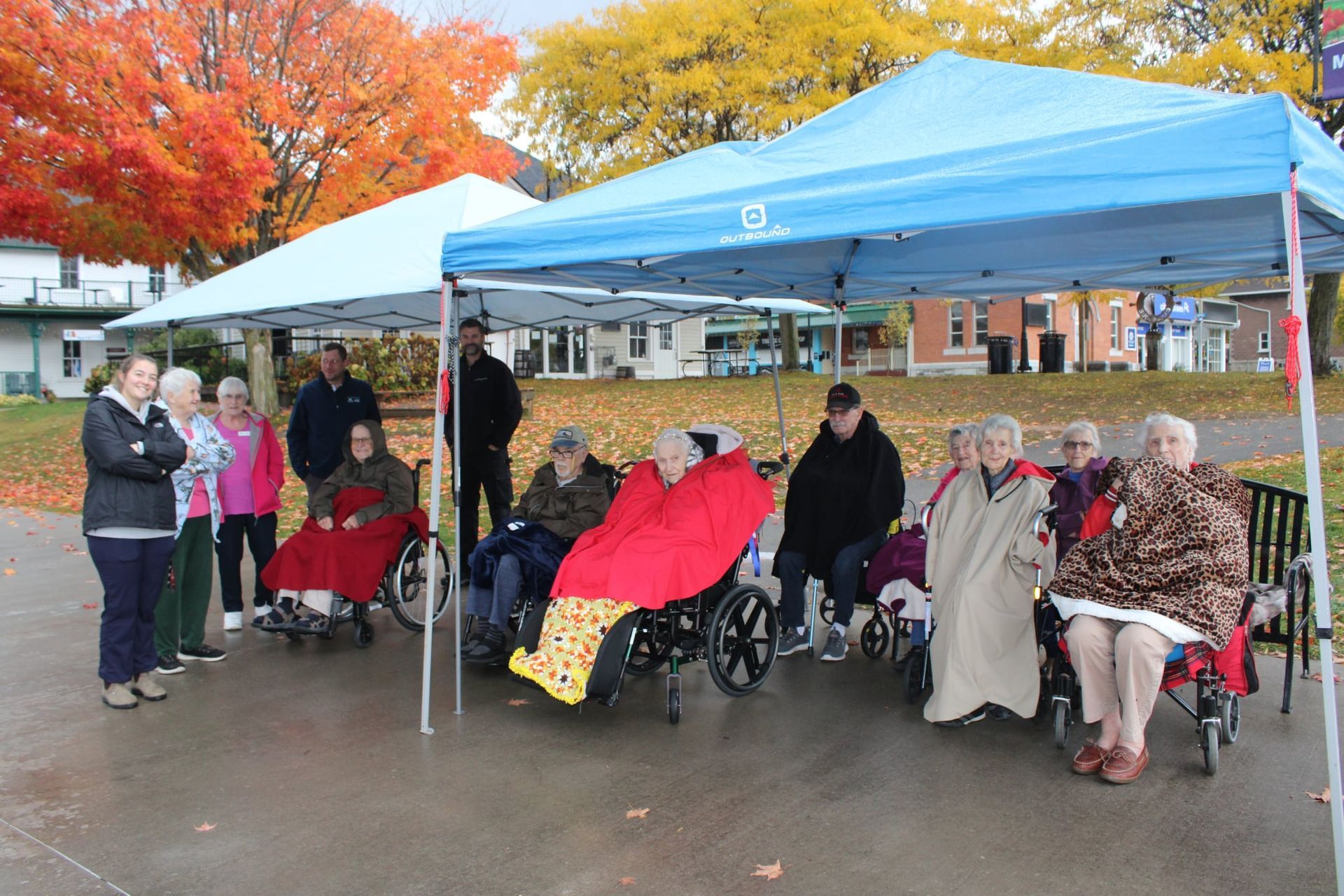 People gathered under blue tents outdoors in autumn, with several wheelchairs and colorful blankets.