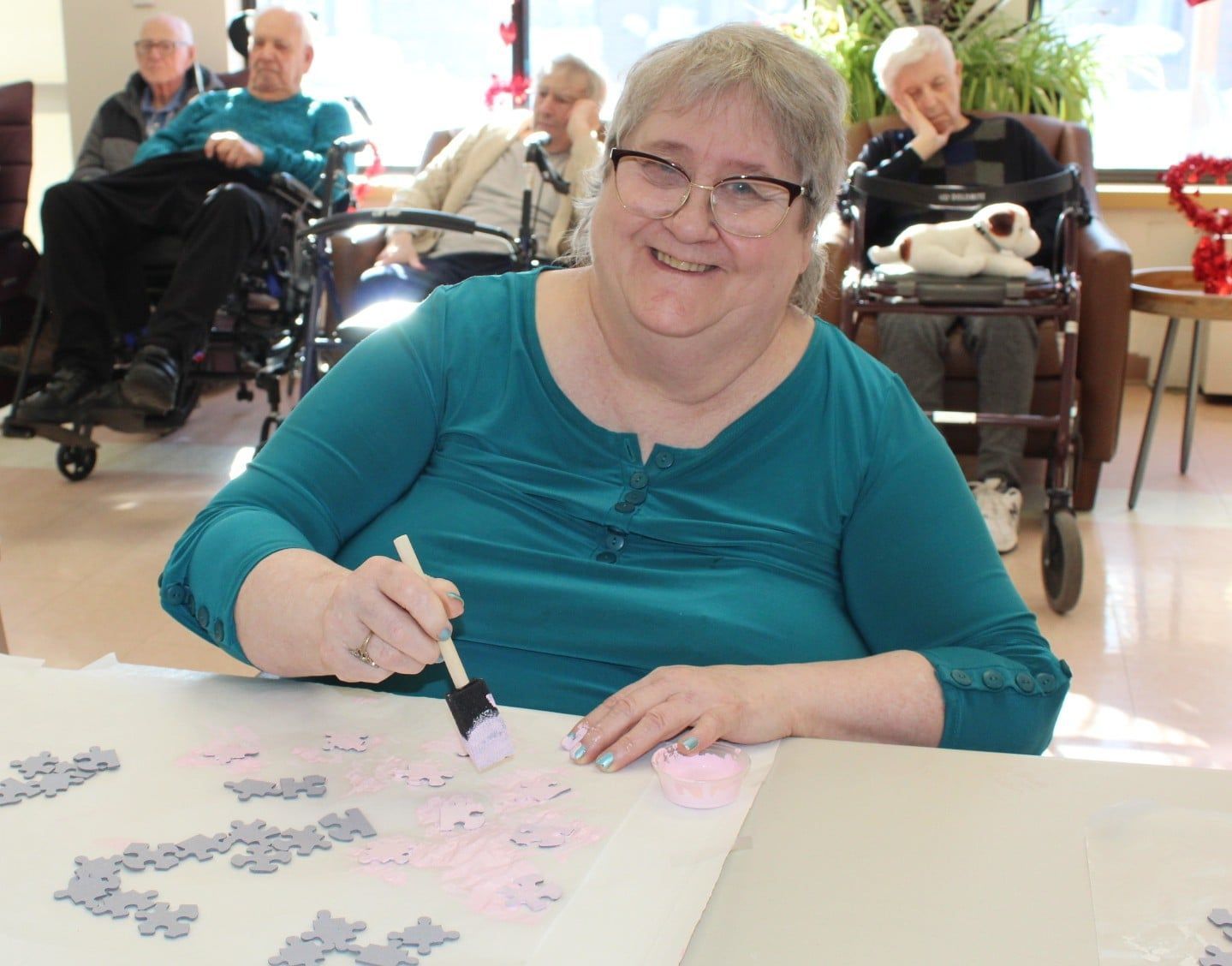 Woman smiling and painting a puzzle at a table in a bright community room, with others seated behind her