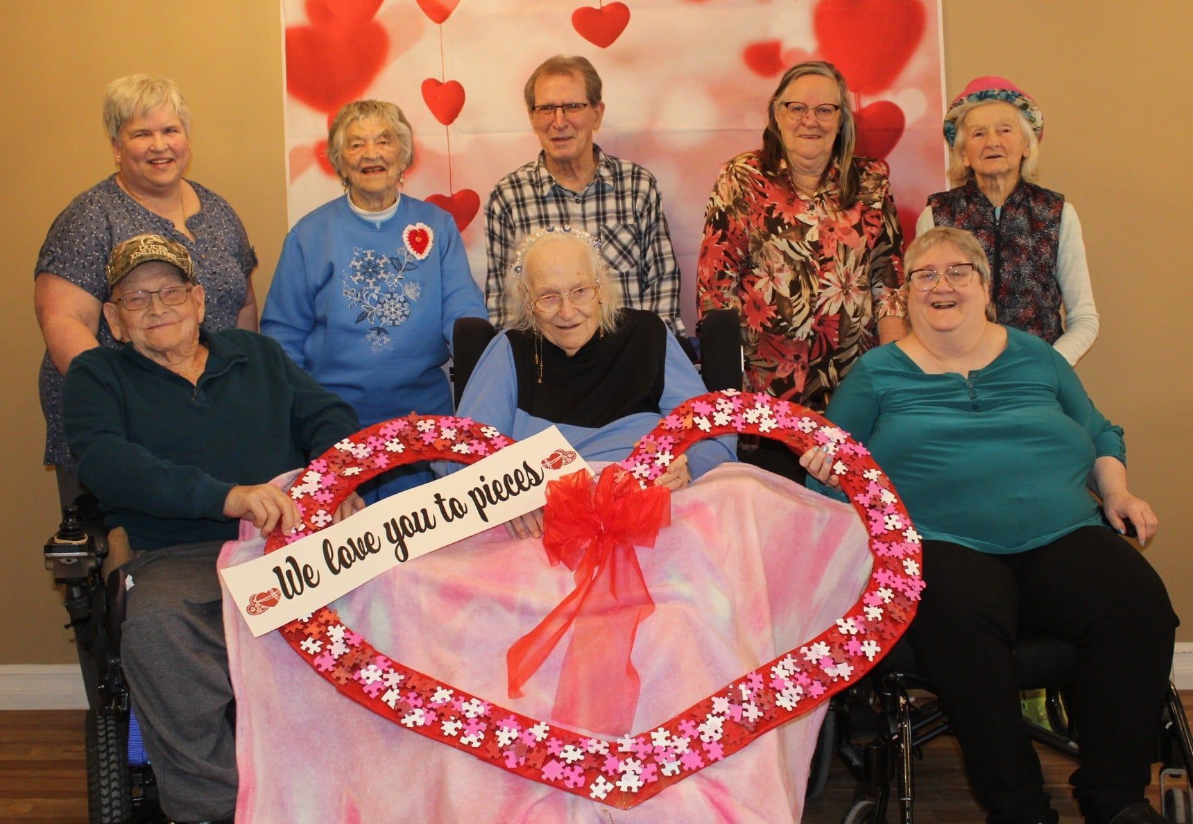 Seven smiling people pose around a heart-shaped Valentine's display with pink and red decorations.